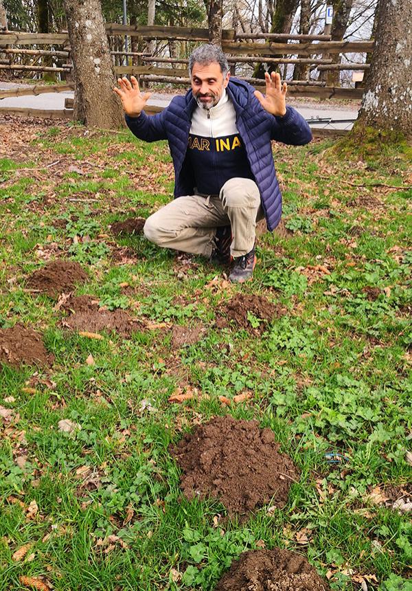 Uomo sorridente inginocchiato su prato, circondato da cumuli di terra, alza le mani felice.